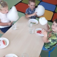 Dzieci siedzą przy stoliku i jedzą watę cukrową i arbuzy.