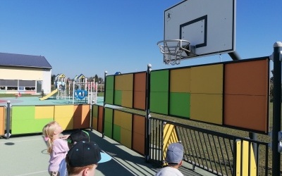 Dzieci grają w koszykówkę na boisku. W tle plac zabaw. 