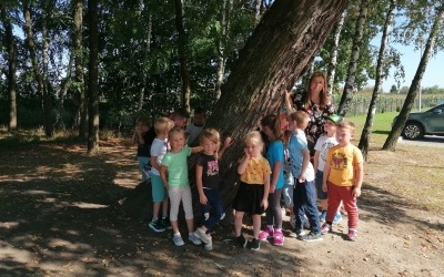 Dzieci oraz pani przytulają się do drzewa. W tle inne drzewa. 