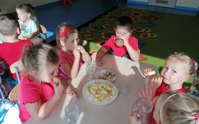Dzieci siedzą przy stoliku. Piją sok i jedzą jabłka. 