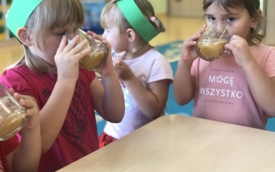 Dziewczynki siedzą przy stoliku, na głowach maja opaski z emblematem jabłka, piją sok jabłkowy.