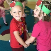 Chłopiec i dziewczynka tańczą w parze. Na głowach mają opaski z emblematem jabłka. W tle tańczą inne dzieci.