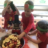 Trzech chłopców wykonuje sok jabłkowy przy pomocy sokowirówki. Chłopcy mają na głowach opaski z emblematem jabłka.