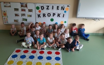 Dzieci siedzą na podłodze pod tablicą z napisem Dzień Kropki a przed nimi duża plansza w kolorowe kropki.