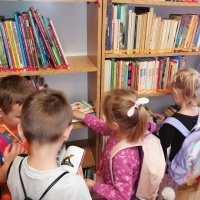 Dzieci z plecakami wypożyczają książki w bibliotece. W tle regały z książkami. 