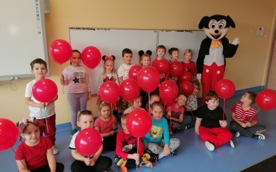 Dzieci z balonami stoją w sali przedszkolnej. W tle tablica i Myszki Miki. 