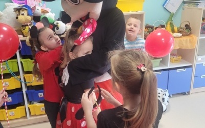 Dwie dziewczynki przytulają Myszkę Miki. W tle widoczne inne dzieci oraz półki z zabawkami. 