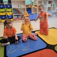 Trzy dziewczynki ubrane na pomarańczowo siedzą na dywanie i przeplatają włóczkę przez otwory zrobione w szablonie dyni. W tle widoczna półka z zabawkami.
