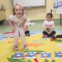 Na zdjęciu widać uśmiechniętą dziewczynkę, która układa ilustracje bałwanków. W tle widać inne dzieci które siedzą na dywanie.