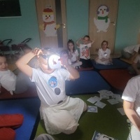 Dzieci siedzą na dywanie. Dziewczynka trzyma w ręce latarkę i podświetla obrazki. 