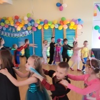 Grupa dzieci tańczy na sali. W tle kolorowa dekoracja z balonami.