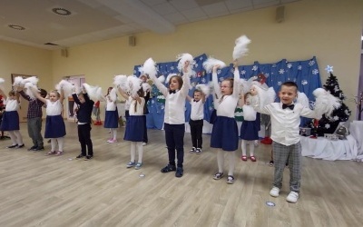Grupa dzieci tańczy na sali z białymi pomponami. W tle dekoracja świąteczna.