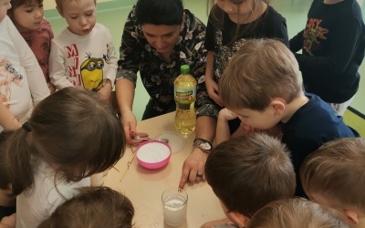 Nauczyciel demonstruje dzieciom doświadczenia z solą, dzieci ustawione wokół stolika obserwują co zachodzi.  