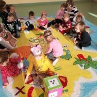 Dzieci wraz z nauczycielkami siedzą na kolorowym dywanie. Dziewczynka w żółtej bluzce trzyma w rękach dużą kolorową kostkę do gry. 