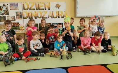 Dzieci siedzą pod tablicą z napisem Dzień Dinozaura i pozują do zdjecia. 