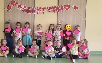 Grupa dzieci pozuje do zdjęcia w rękach trzymają czerwone serduszka. W tle napis Walentynki.