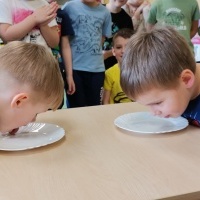 Dwaj chłopcy pochyleni nad talerzykami z mlekiem a reszta grupy obserwuje co robią. 
