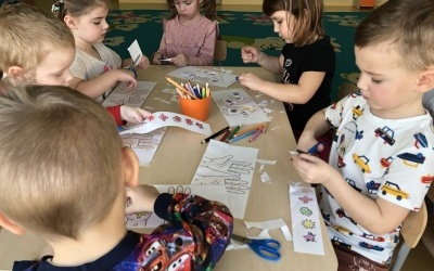 Grupa dzieci siedzi przy stoliku, wykonują pracę plastyczną. Wycinają emblematy bakterii.