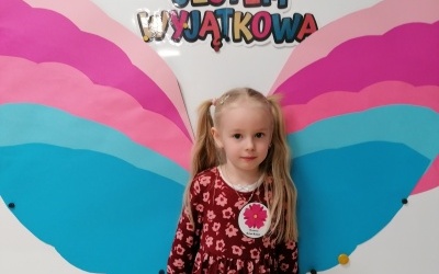 Dziewczynka stoi przed tablicą. W tle napis jestem wyjątkowa oraz kolorowe skrzydełka. 