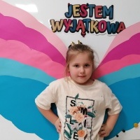 Dziewczynka stoi przed tablicą. W tle napis jestem wyjątkowa oraz kolorowe skrzydełka. 