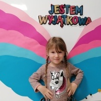 Dziewczynka stoi przed tablicą. W tle napis jestem wyjątkowa oraz kolorowe skrzydełka. 