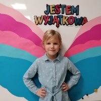 Dziewczynka stoi przed tablicą. W tle napis jestem wyjątkowa oraz kolorowe skrzydełka. 