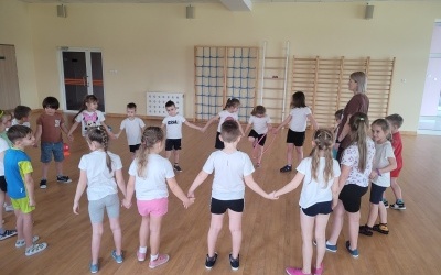 Grupa dzieci w strojach sportowych wykonuje ćwiczenia na sali gimnastycznej.