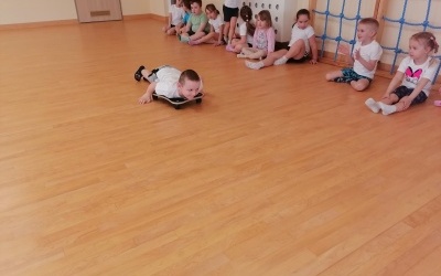 Dzieci ćwiczą na sali gimnastcznej. Chłopiec jedzie na deseczce. 