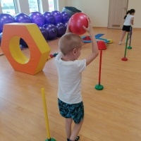 Dzieci ćwiczą na sali gimnastcznej. Chłopiec z piłką pokonuje tor przeszkód. 