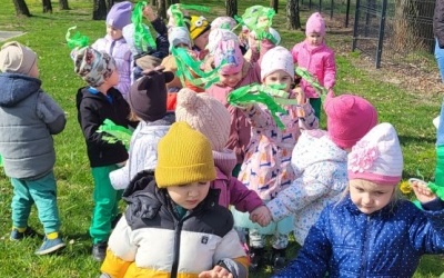 Grupa dzieci idzie w wiosennym pochodzie. W rękach trzymają zielone wstążki.
