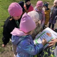 Grupa dzieci na spacerze szuka oznak wiosny. Dziewczynka zaznacza znalezioną oznakę wiosny na karcie obserwacji. 