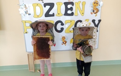 Chłopczyk i dziewczynka pozują  do zdjęcia na tle tablicy z napisem Dzień Pszczoły. Na głowach mają pszczele nakrycie głowy, w rękach trzymają akcesoria.a