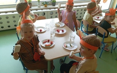Dzieci siedzą przy stoliku i zajadają smakołyki: truskawki, arbuzy i lody.