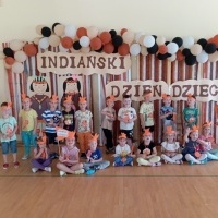 Dzieci stoją ustawione na sali gimnastycznej na ściance z napisem Indiański Dzień Dziecka