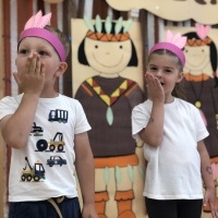 Dziewczynka i chłopiec stoją na sali gimnastycznej. Na głowach mają pióropusze indiańskie. W tle dekoracja indiańska.