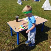 Chłopczyk ubrany w niebieska bluzkę i zielony pióropusz, maluje farbami.